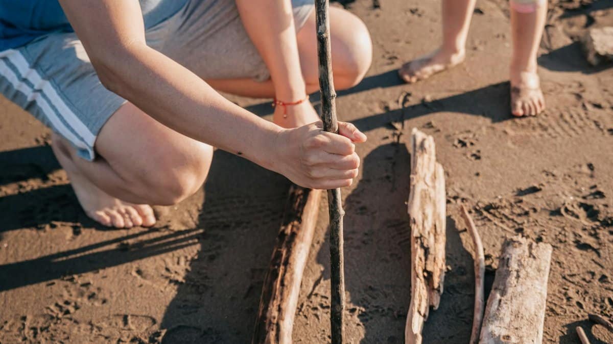 Hands holding a stick, crafting on a sunny beach with driftwood.