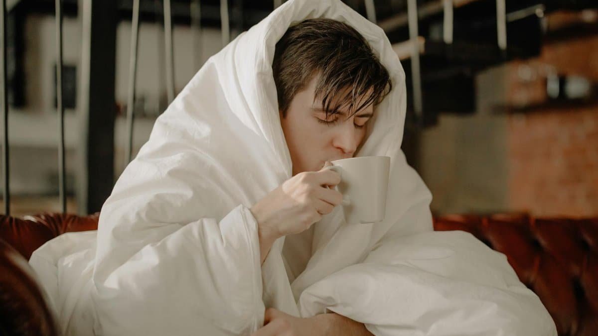 Young man wrapped in blanket sipping hot beverage at home, embracing comfort and relaxation.