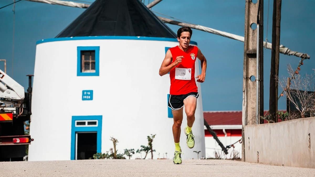 A fit young man in a red jersey jogs energetically near a traditional windmill.