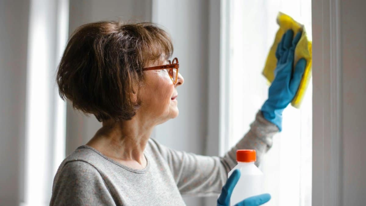 Elderly woman cleansing window indoors using gloves and cleaner.