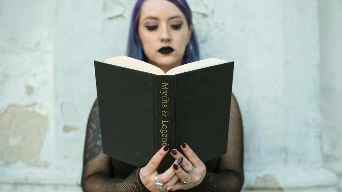 Caucasian woman with gothic style reads 'Myths & Legends' book outside.