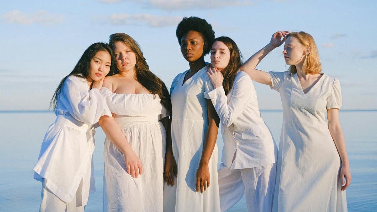 A diverse group of women in white outfits stand together by the ocean, celebrating unity and sisterhood.