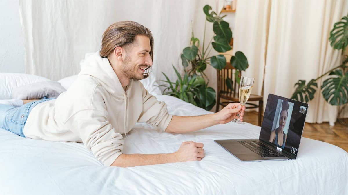 A man smiles while video calling and holding a glass of wine in a cozy bedroom setting.