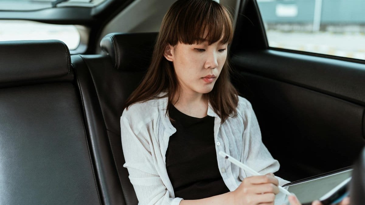 Concentrated Asian female in casual clothes working on modern tablet with stylus while riding in car backseat
