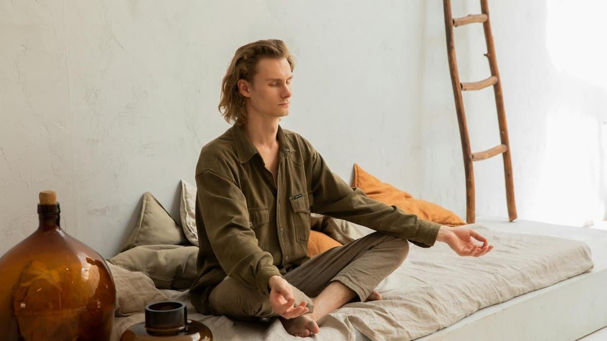 Young mindful barefoot male with closed eyes sitting in Padmasana pose while practicing yoga at home