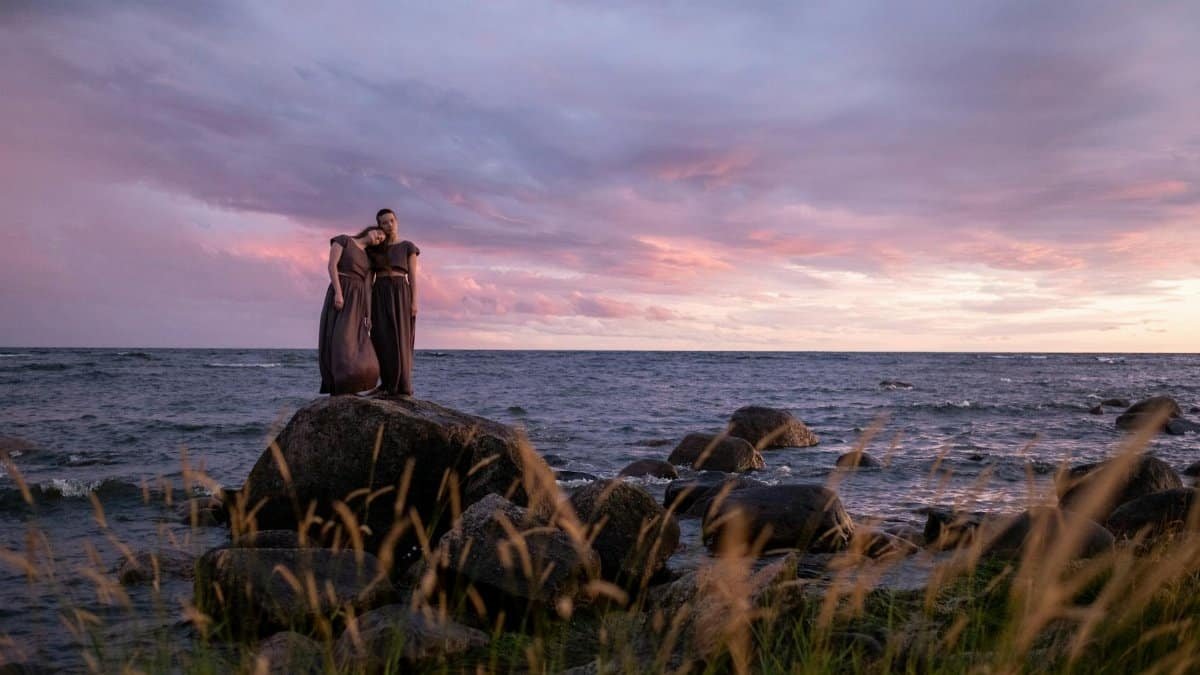 Two women stand on rocks by the sea at sunset, embracing under a colorful sky.