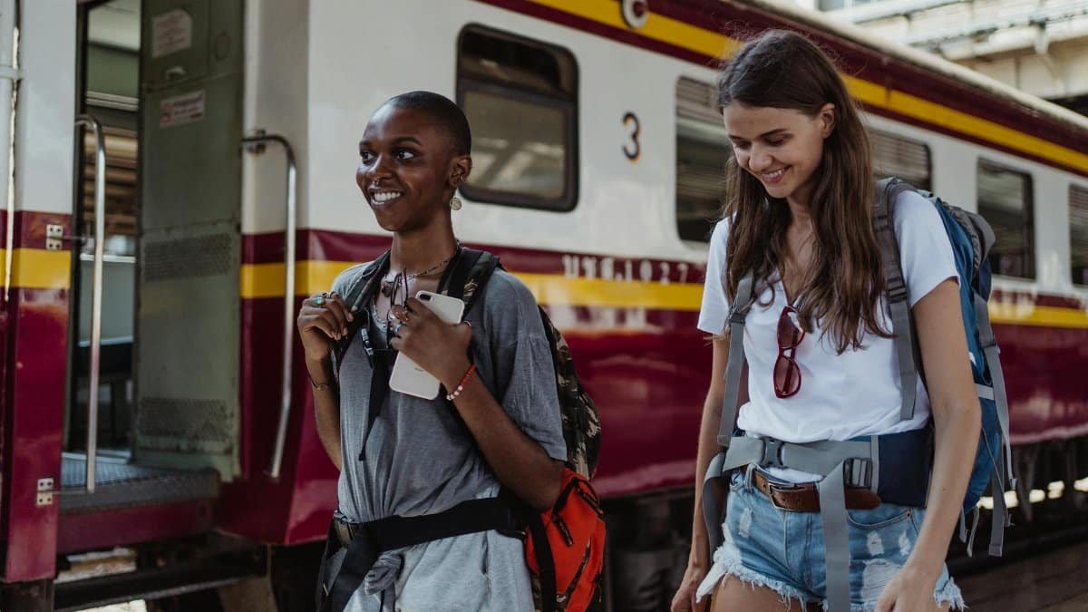 Two women with backpacks enjoying a train journey, smiling and relaxed, standing next to a train.
