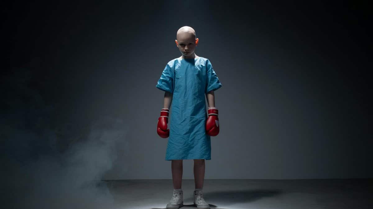 A young child in a hospital gown wears boxing gloves, symbolizing strength and resilience.
