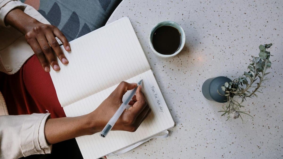 Person writing in a planner with coffee at a café, focusing on planning and productivity.
