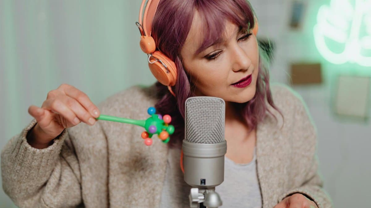 A woman records ASMR with a microphone and headphones indoors, showcasing studio equipment.
