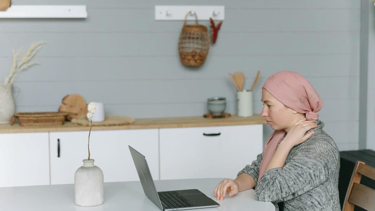 Woman with head wrap sitting at a kitchen table, using a laptop, symbolizing resilience.