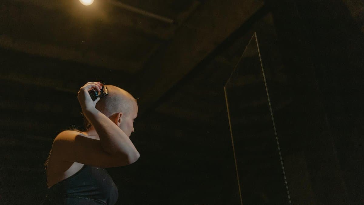 A woman shaves her head in a dimly lit room, reflecting empowerment and change.