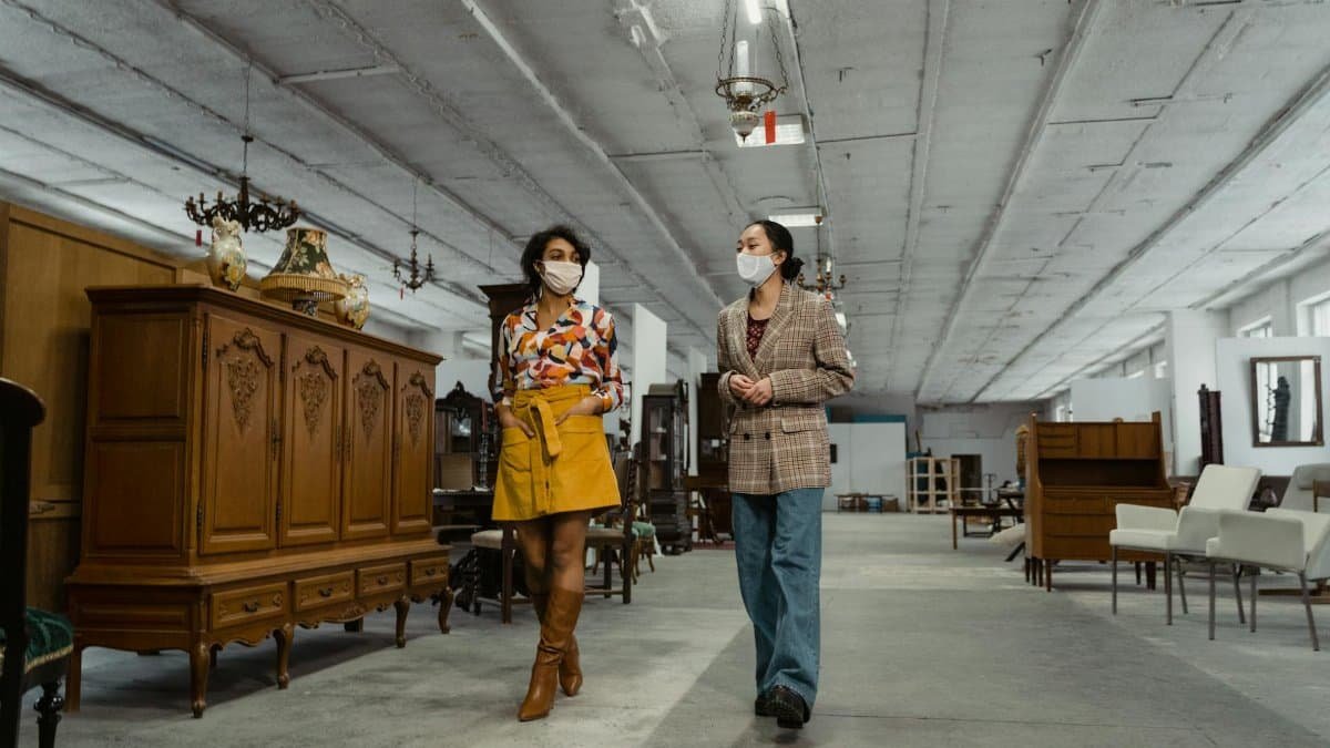 Two women wearing masks explore a vintage furniture store, highlighting fashion and the new normal.