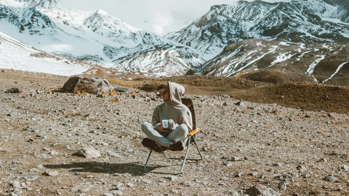 Full body of man sitting on folding chair and drinking tea with high mountains covered with snow on background