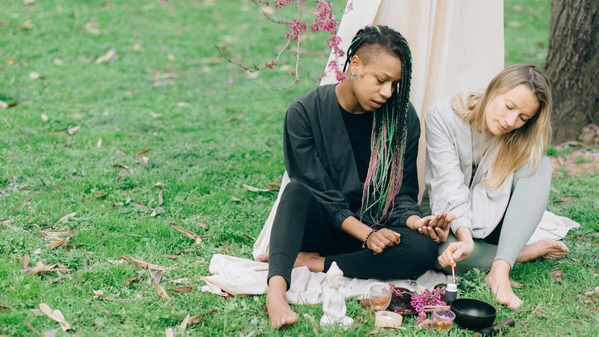 Two women meditating outdoors with yoga accessories, embracing relaxation and diversity.
