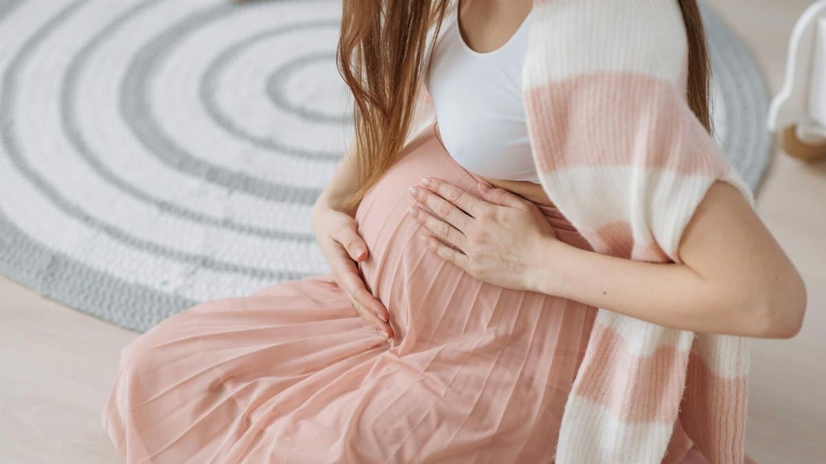Pregnant woman sitting indoors, tenderly holding her baby bump in soft, cozy attire.