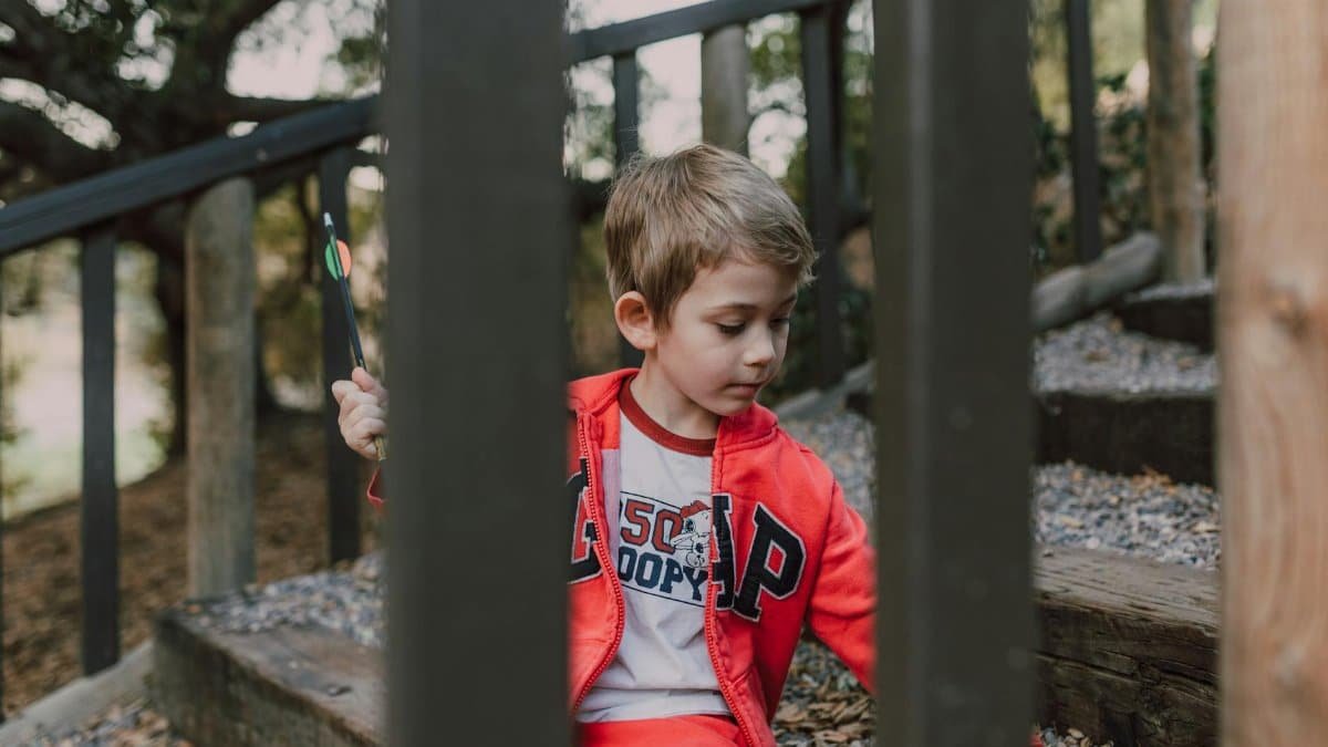 A young boy holding an arrow sits on wooden outdoor steps surrounded by trees.