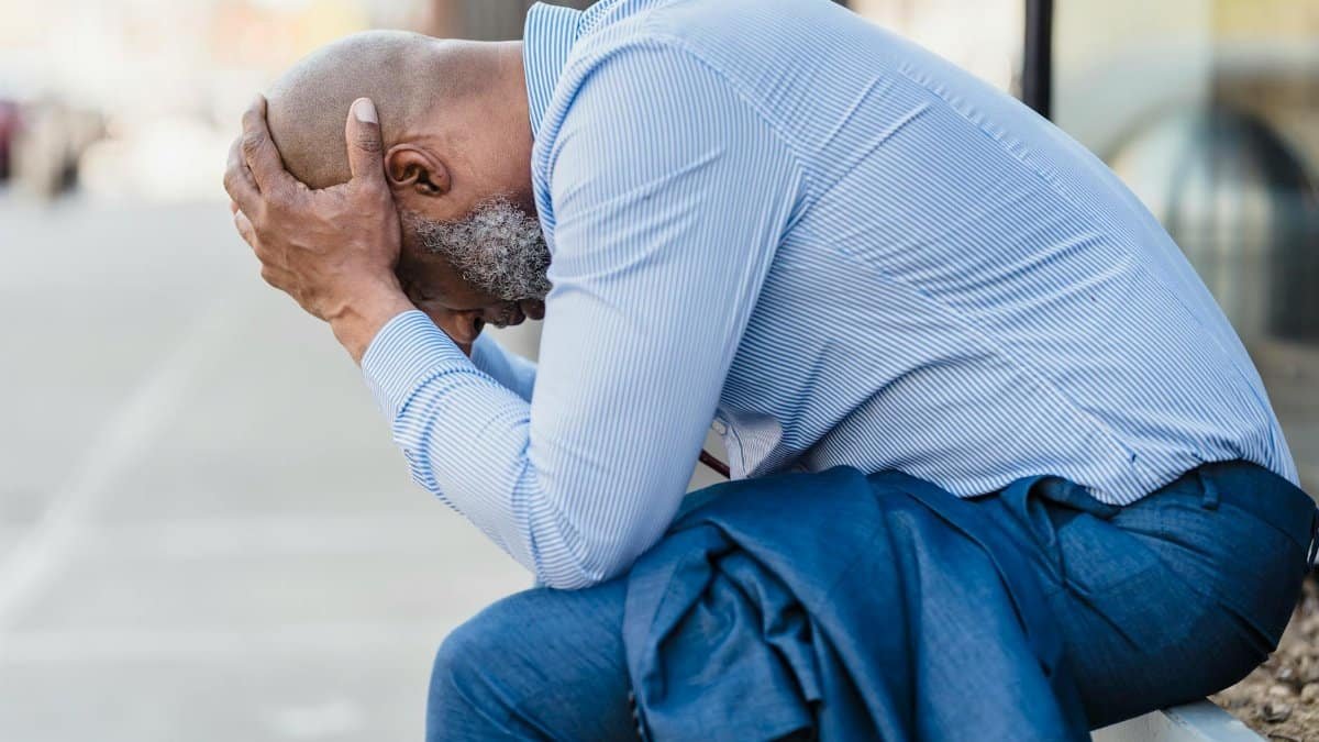 An adult man sitting outdoors, showing stress and contemplation with his head in hands.