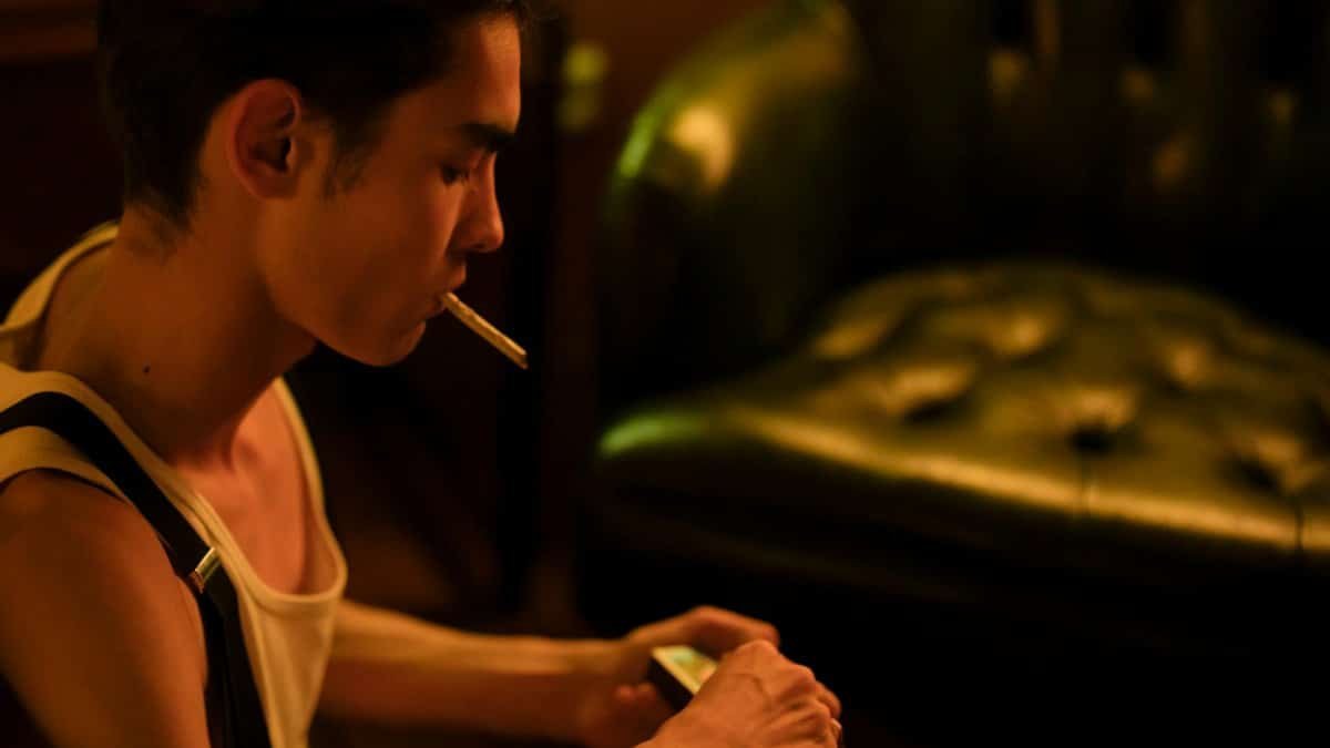 A young man sits indoors, smoking a cigarette in a dimly lit room.