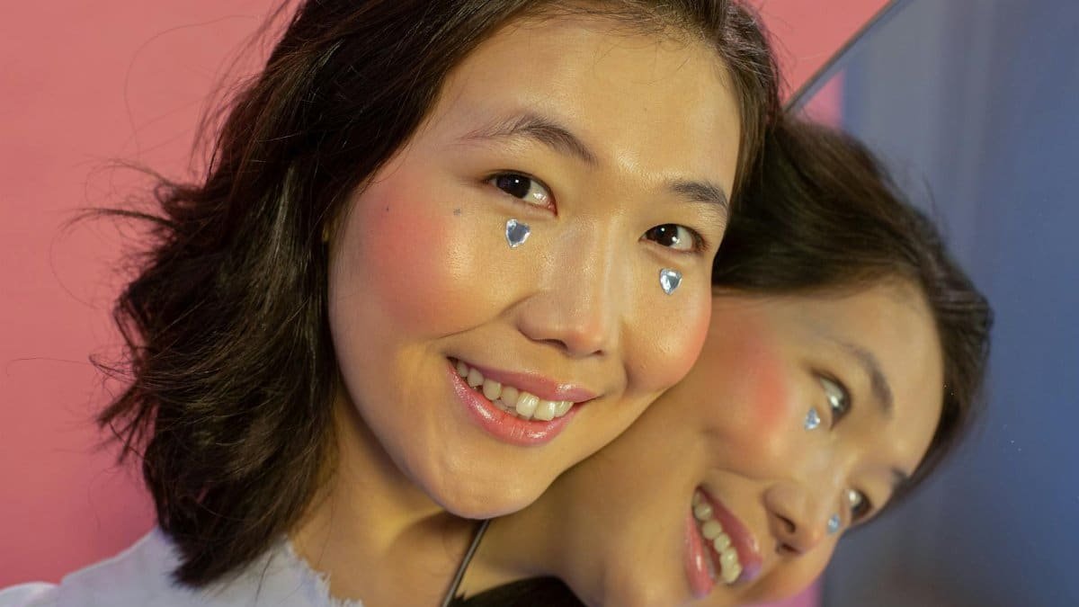 Smiling young ethnic lady in makeup looking at camera with shining rhinestones in shape of hearts on cheeks near mirror with self reflection on pink background in light studio