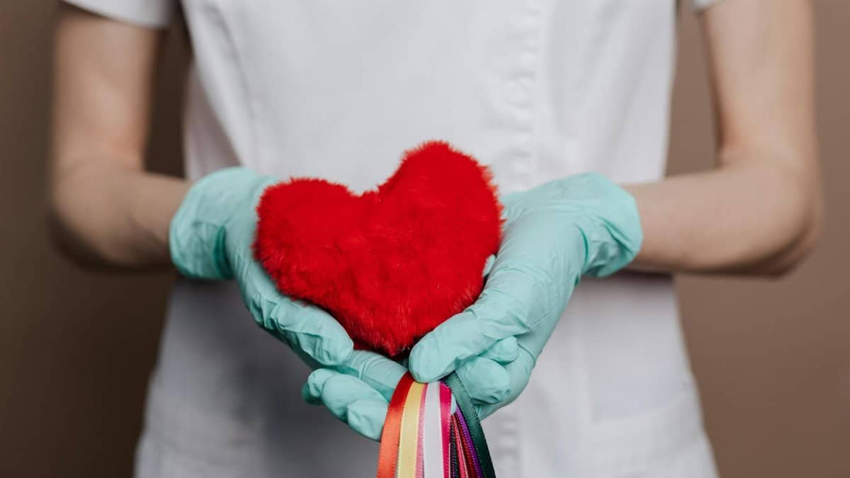 A nurse in gloves holds a red heart with rainbow strings, symbolizing love and healthcare.