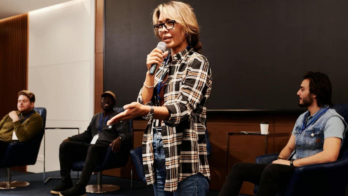 A confident woman speaks into a microphone during a panel discussion indoors.