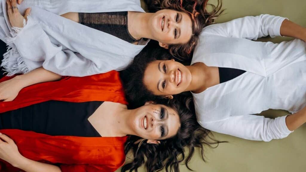 Three smiling women lying on the floor wearing vibrant clothing, symbolizing joy and friendship.