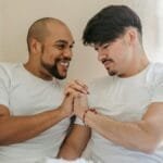 Two men sharing a moment of affection on a bed, holding hands and smiling naturally.