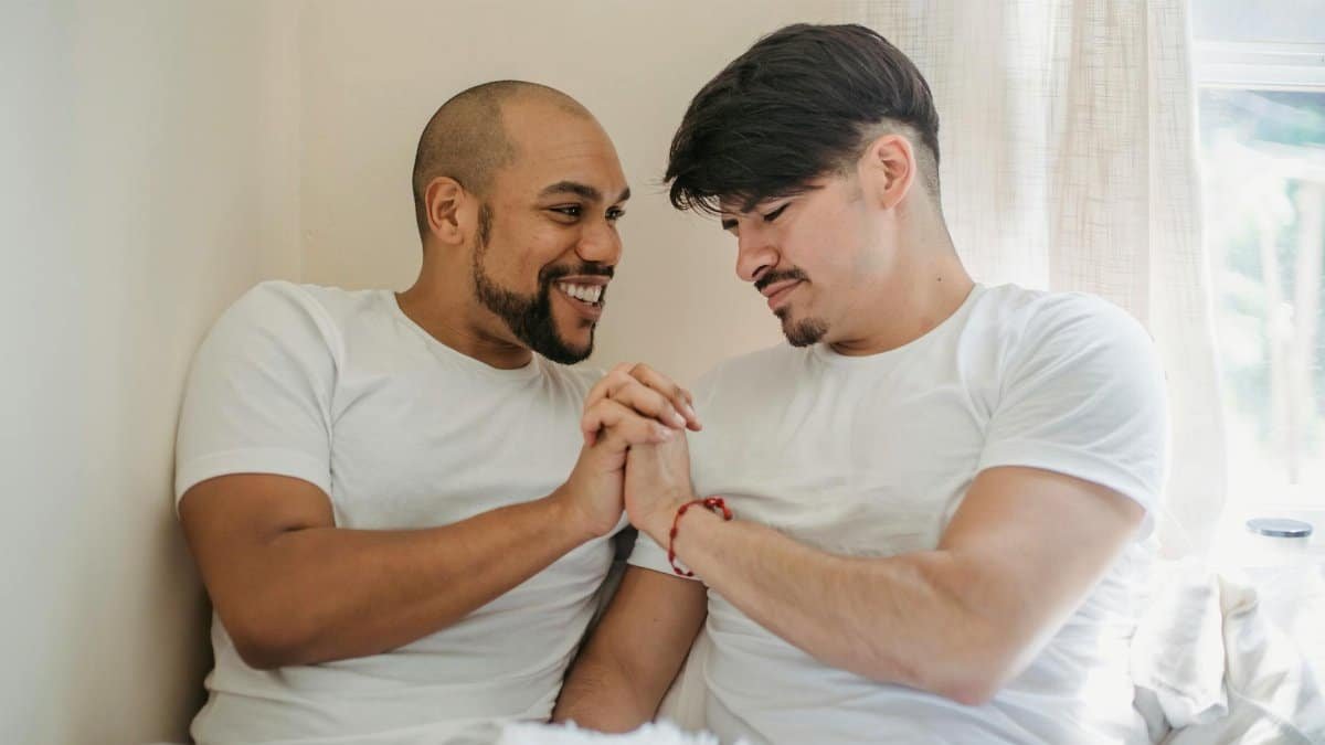 Two men sharing a moment of affection on a bed, holding hands and smiling naturally.