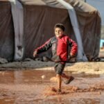 A joyful young boy runs through a muddy puddle in a refugee camp in Idlib, Syria, encapsulating resilience and innocence.