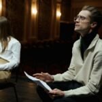 Two actors rehearsing with scripts in a dimly lit theater auditorium.