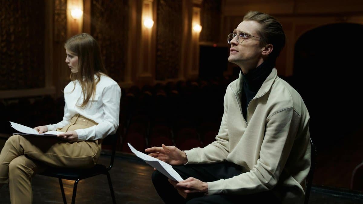 Two actors rehearsing with scripts in a dimly lit theater auditorium.