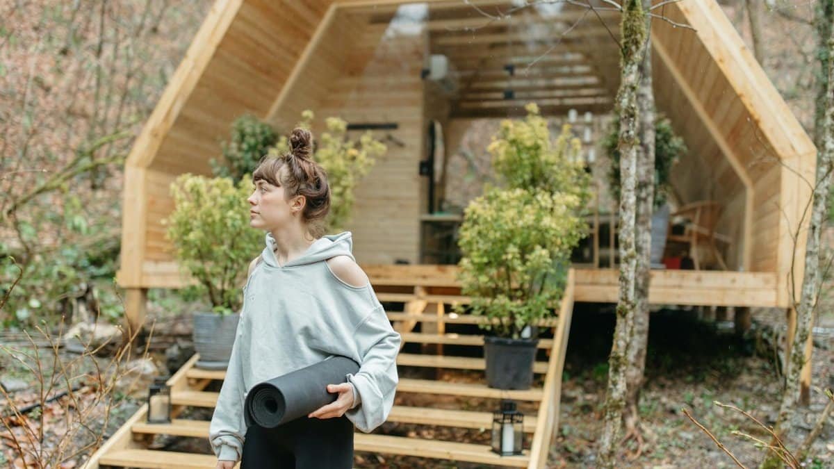 Woman holding a yoga mat outside a wooden cabin in a forest, ready for a wellness retreat.