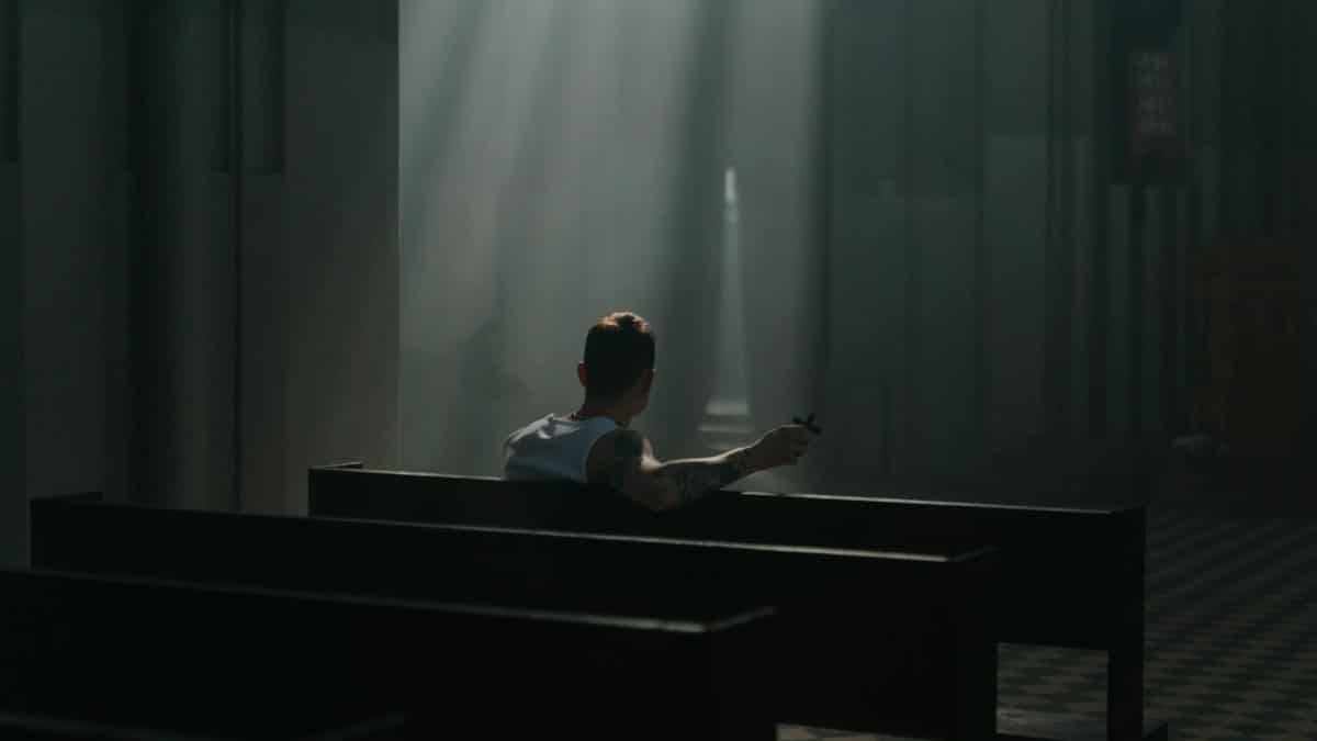 Tattooed man sitting in church pews under a hazy beam of light, holding a crucifix.