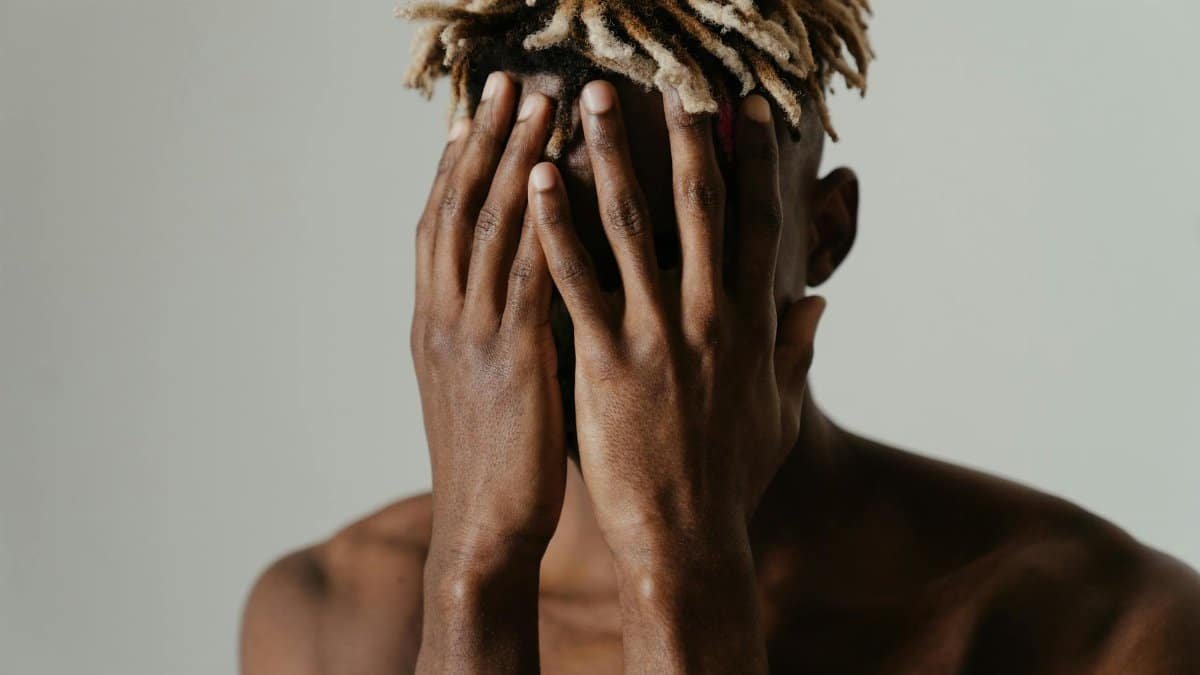 A shirtless young man with dreadlocks covers his face in a contemplative pose, expressing emotion.