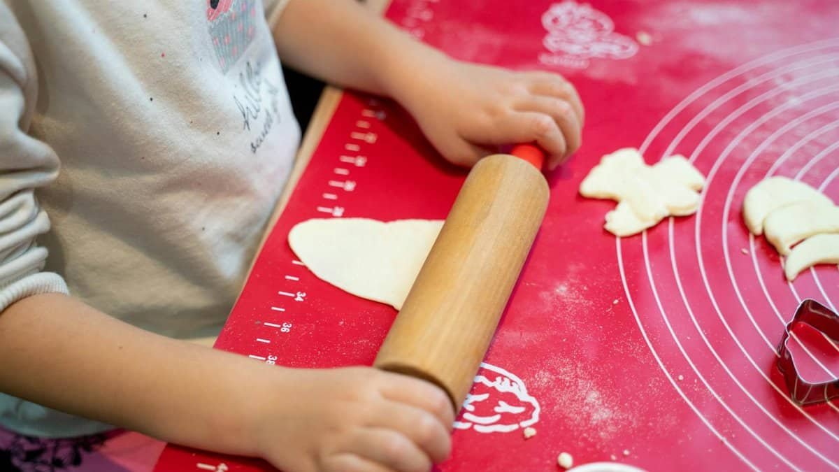 Young child rolling dough on a red baking mat with cookie cutters.