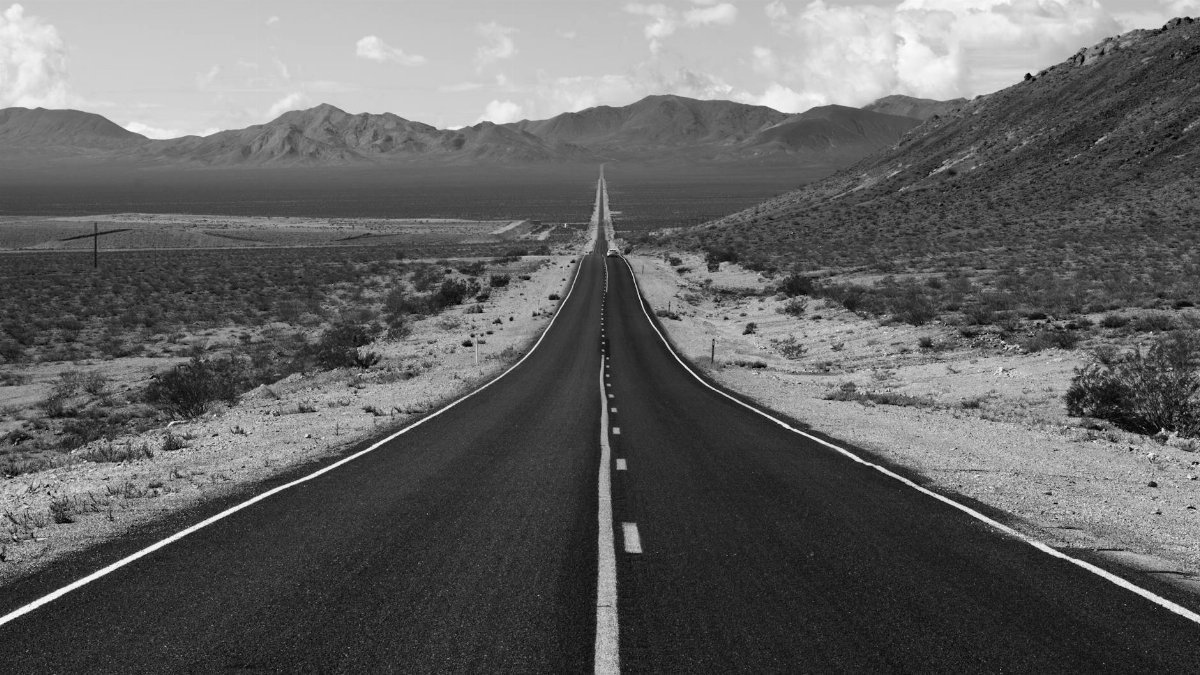Black and white view of a long, empty highway stretching into distant desert mountains.