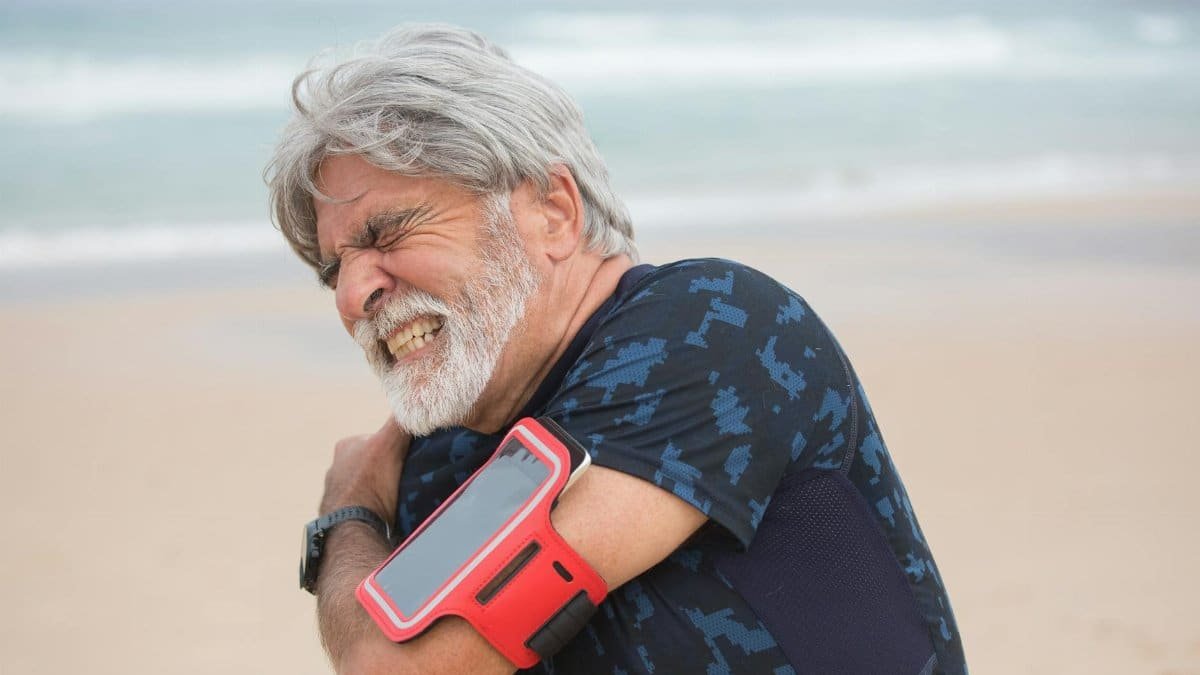 Elderly man grimacing from shoulder pain during seaside walk, showcasing health issue outdoors.