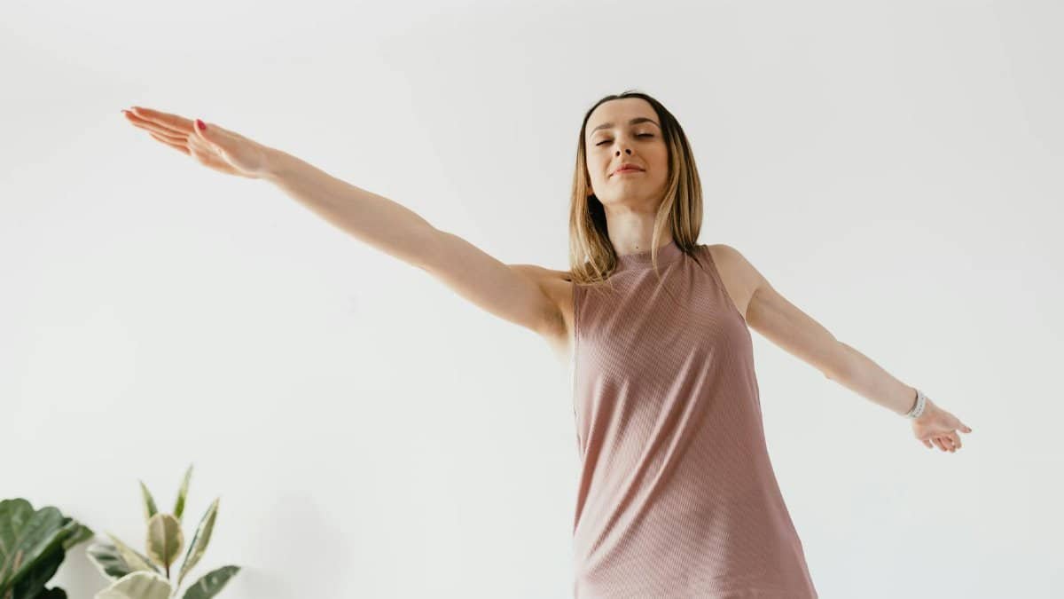 From below of calm young female performing Warrior II yoga asana with extended arms and closed eyes
