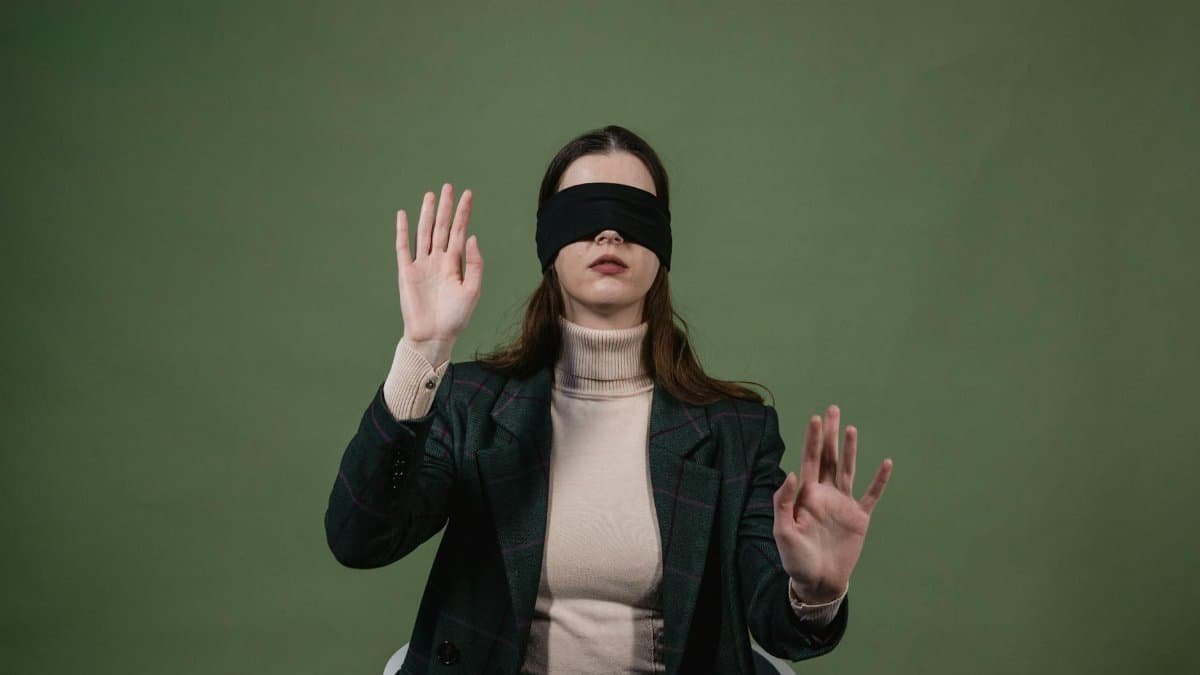 Portrait of a woman blindfolded with raised hands against a green background suggesting sensory exploration.