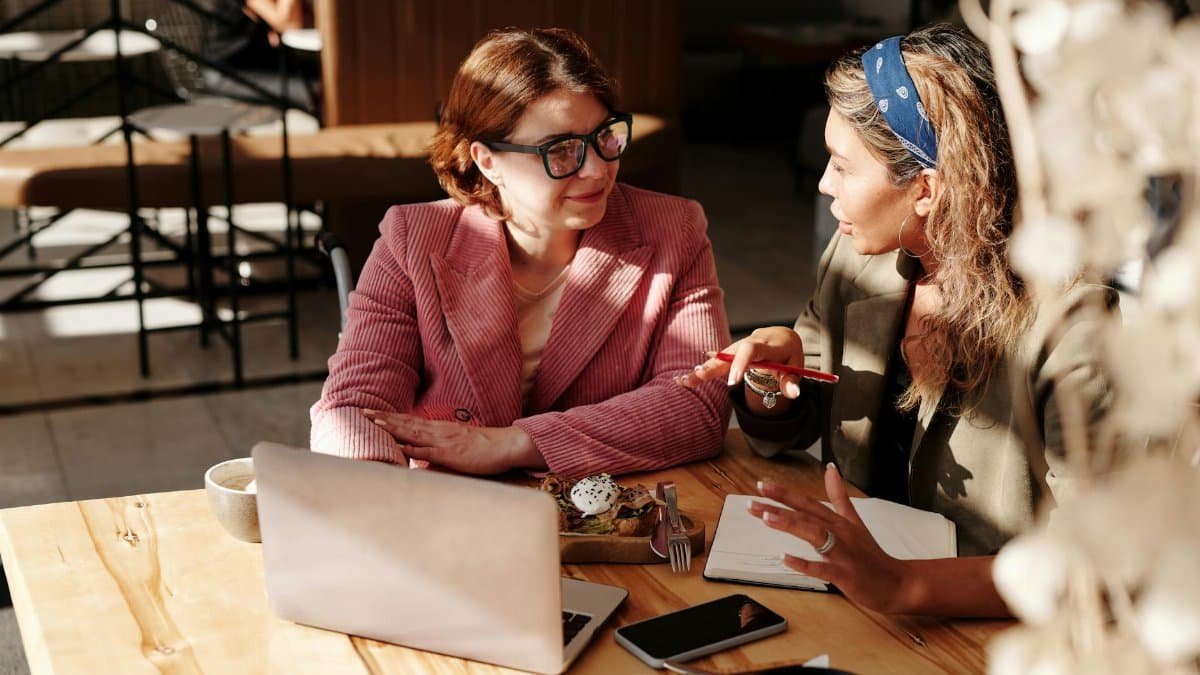 Two businesswomen discussing work at a cafe table with laptops and snacks in sunlight.