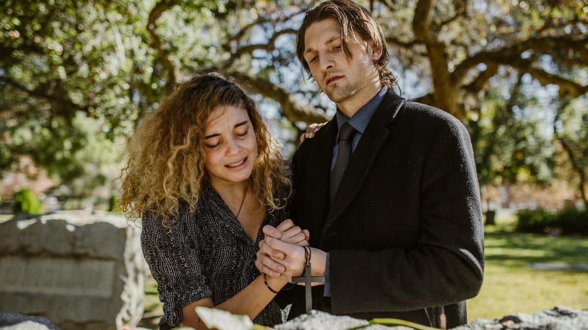 A couple mourns together, holding hands at a grave in a serene cemetery setting.