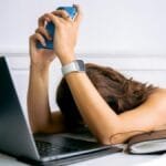 A woman overwhelmed by work, resting head on table with laptop, phone, and smartwatch.