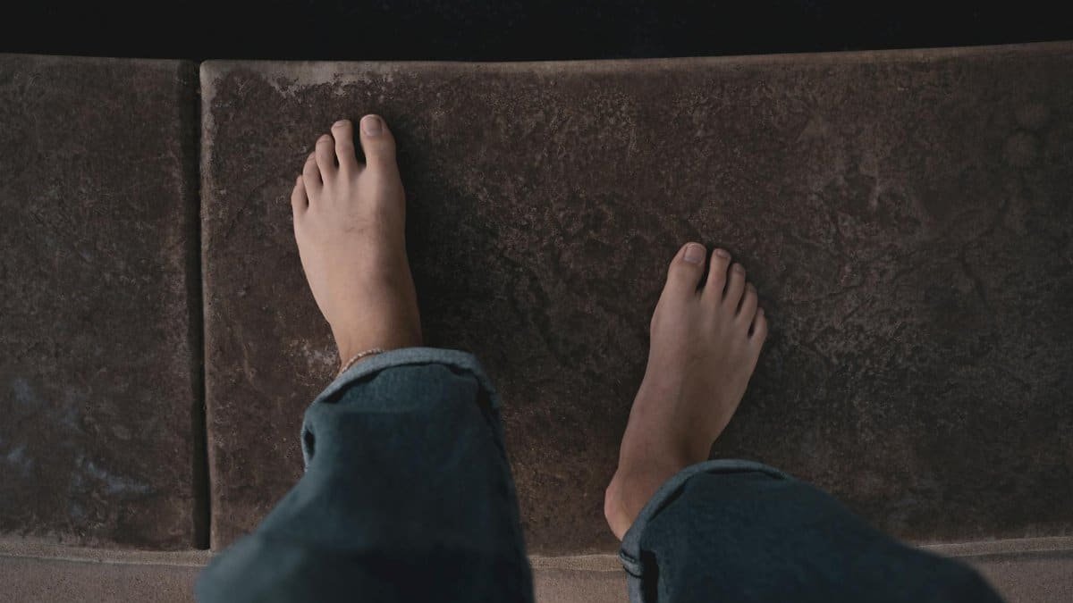 A person's bare feet stand on textured concrete in a first-person view.