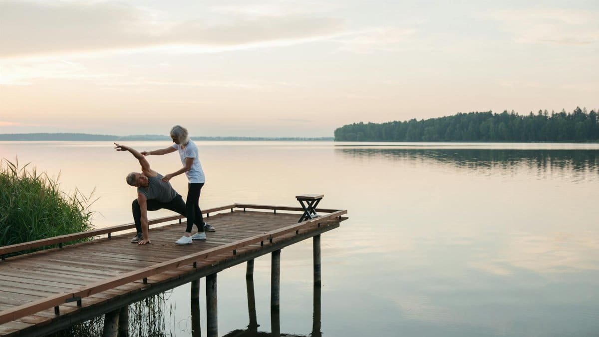 Senior adults practicing yoga on a wooden dock by a serene lake during sunrise, promoting health and mindfulness.