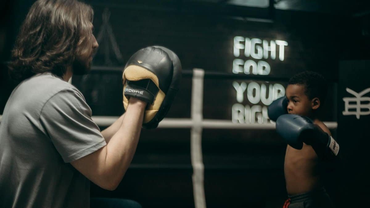 A young boy in boxing gloves trains with his coach in a gym, sign reads 'Fight For Your Right'.