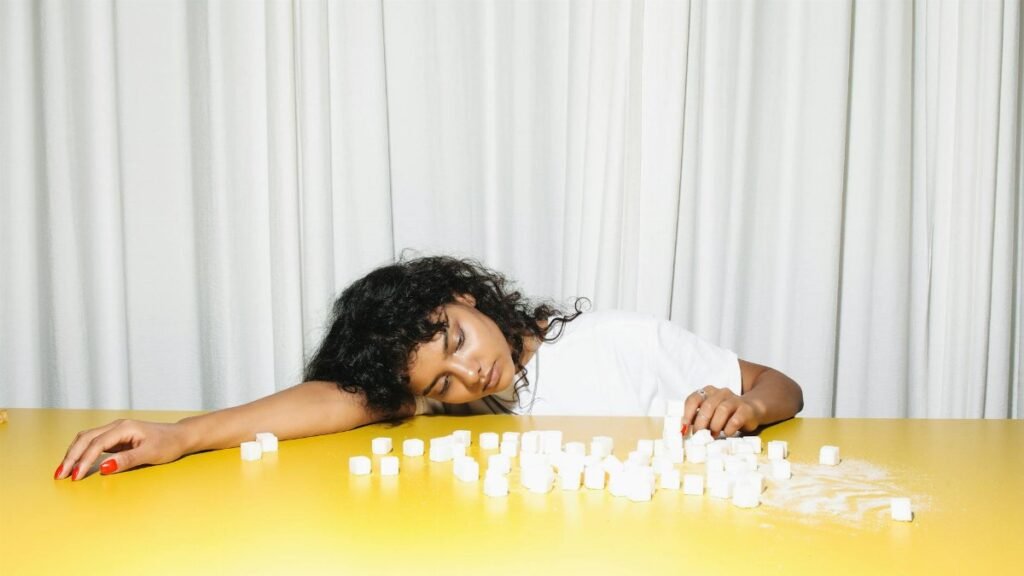 A woman leans on a yellow table surrounded by sugar cubes, highlighting a concept of fatigue and health awareness.