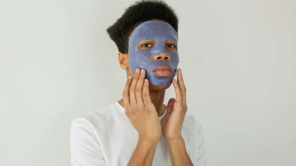 Serious young African American male with moisturizing facial mask applied on face looking away while standing on white background in studio