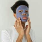 Serious young African American male with moisturizing facial mask applied on face looking away while standing on white background in studio