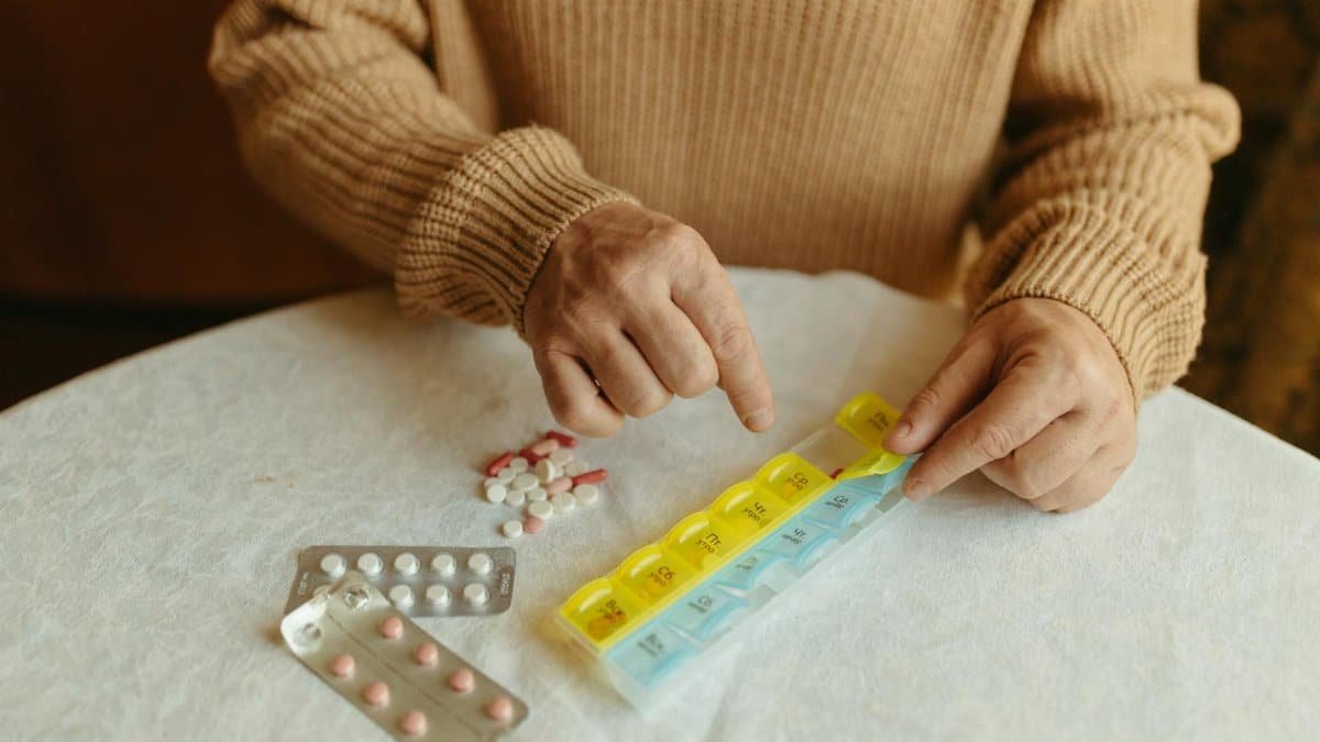 Senior adult organizing daily medications using a weekly pill organizer on a table indoors.